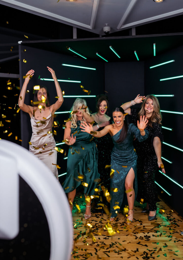 Bridesmaids in green dresses dancing at a photo booth with a neon light backdrop