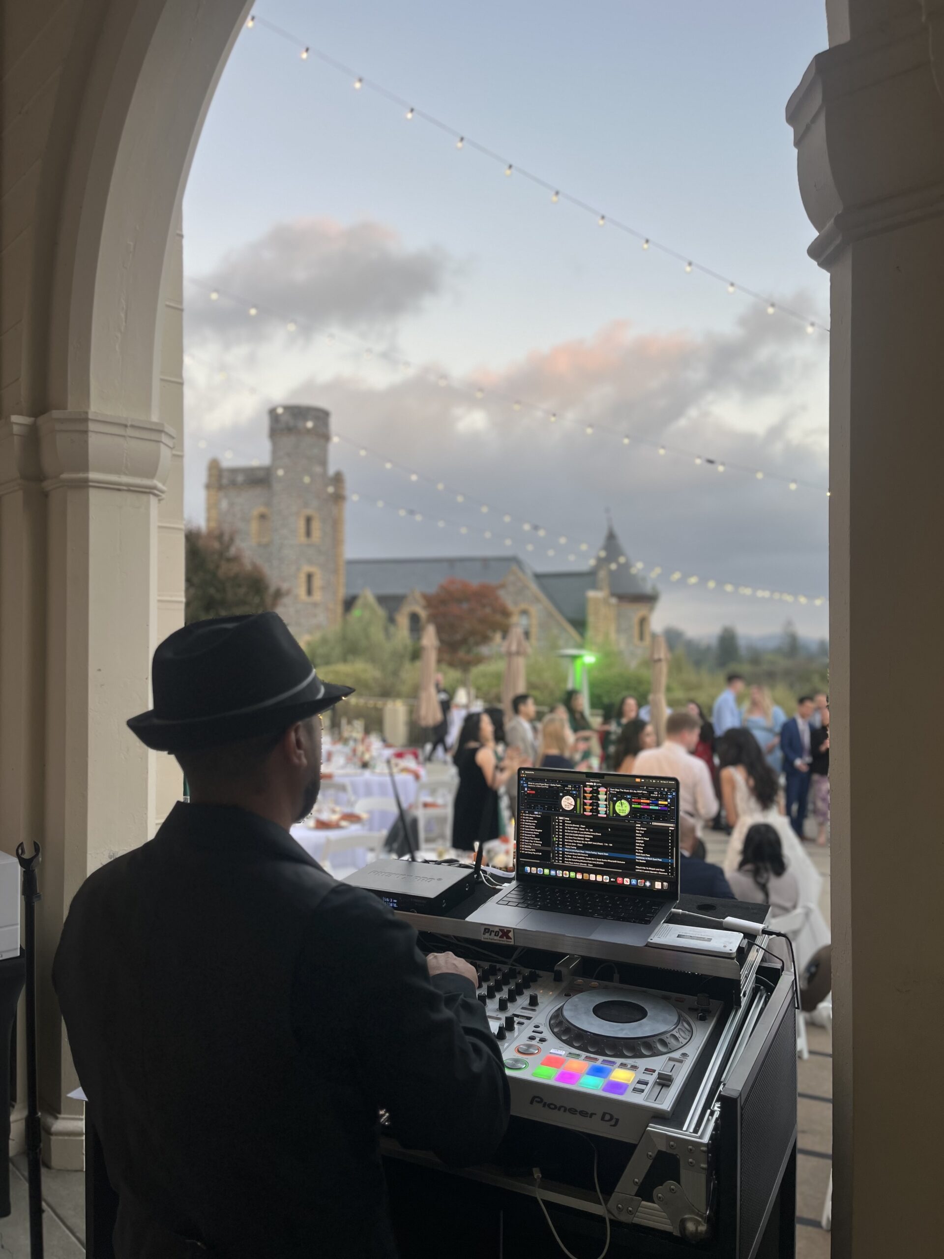 DJ in a black hat performing at an outdoor venue at dusk as guests gather on the lawn