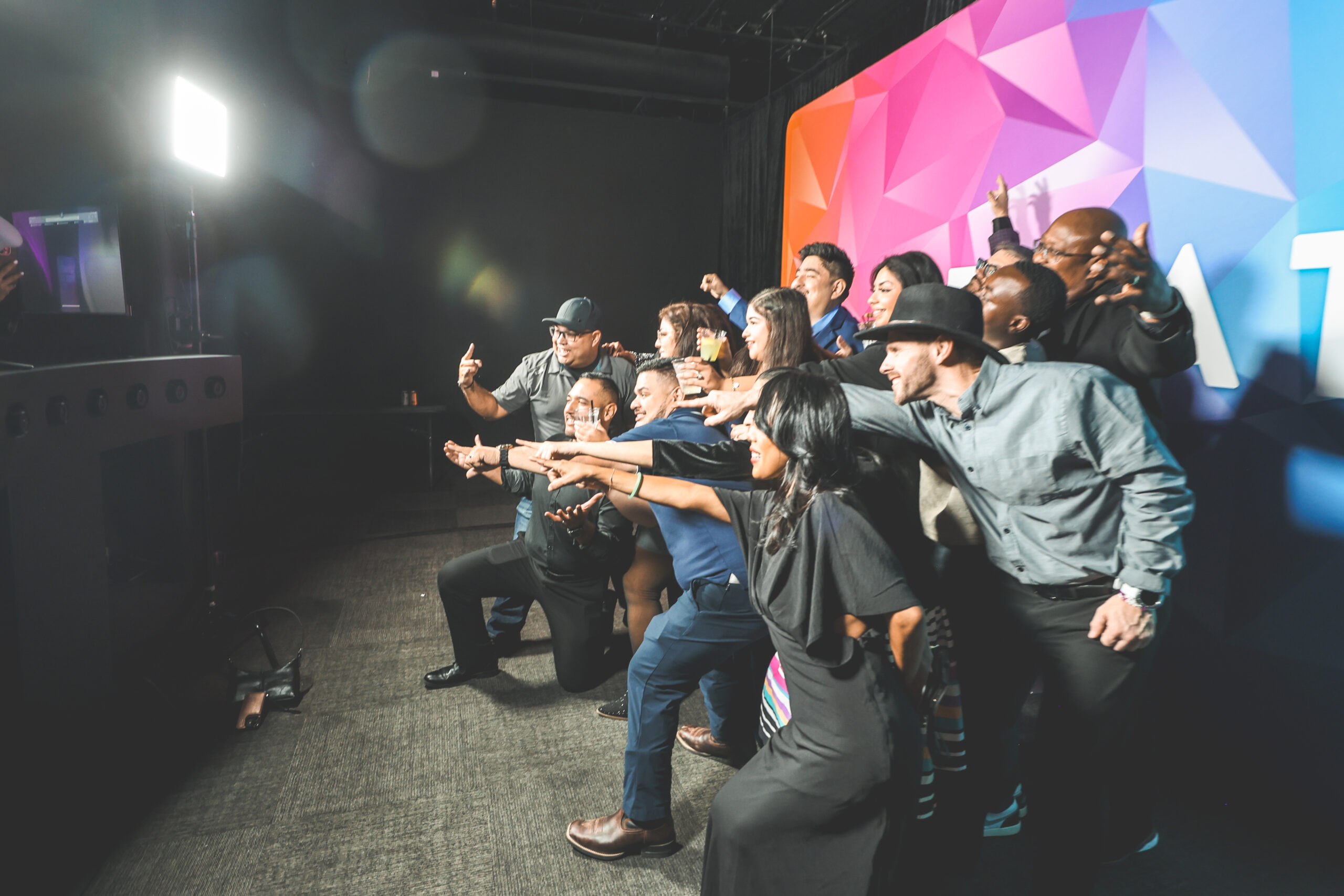 Large group of guests lunging and pointing toward the camera at a photo booth station with a live preview monitor, in front of a colorful CREATE branded backdrop
